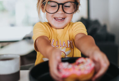 Ein Kind mit Brille greift nach einem bunten Donut auf einem Teller.