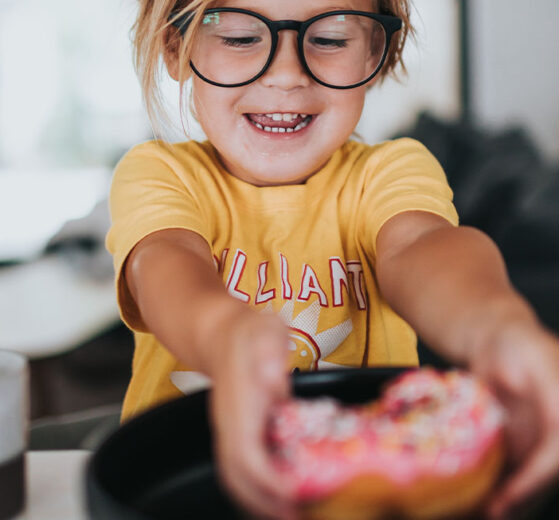 Ein Kind mit Brille greift nach einem bunten Donut auf einem Teller.