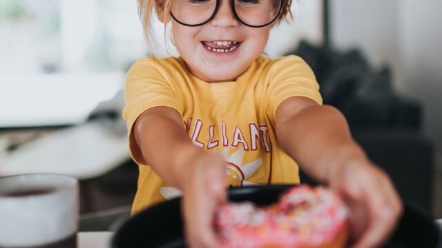 Ein Kind mit Brille greift nach einem bunten Donut auf einem Teller.