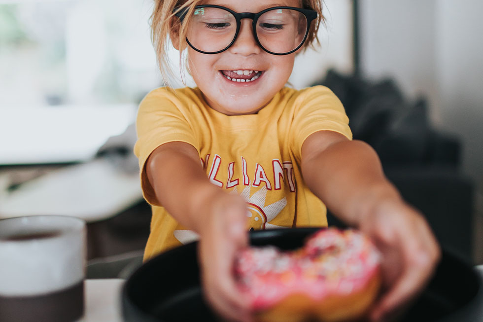 Ein Kind mit Brille greift nach einem bunten Donut auf einem Teller.